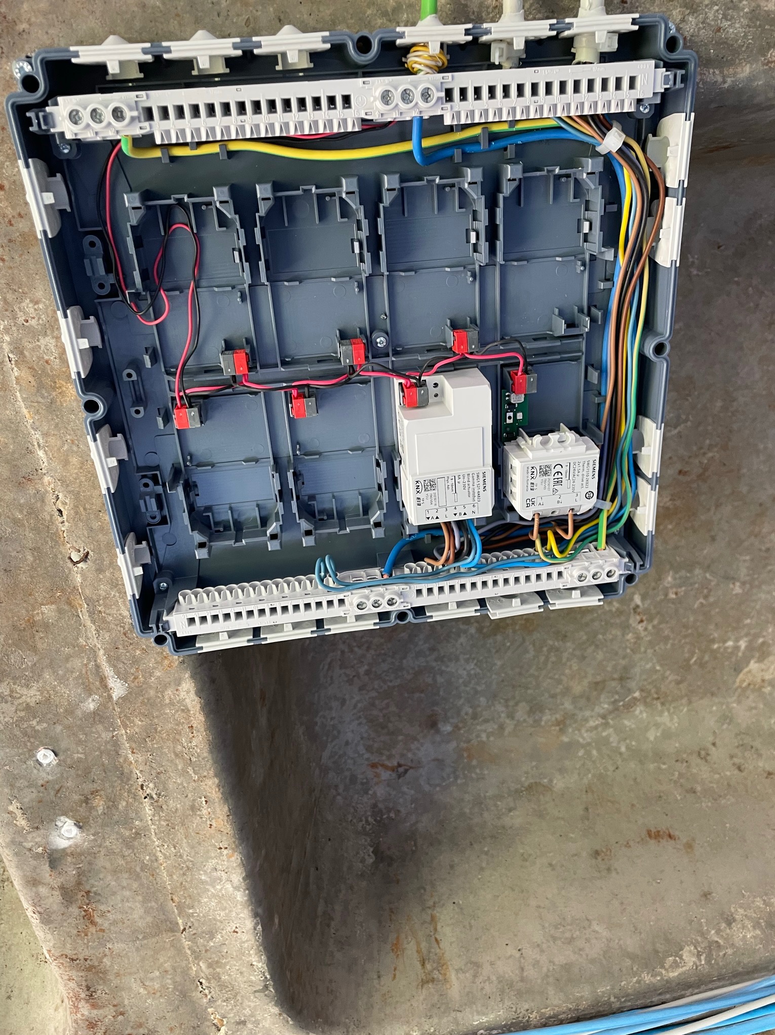 Otwarta, szara skrzynka elektryczna z zamontowanymi modułami Siemens KNX E13, widoczne liczne przewody w różnych kolorach podłączone do listew zaciskowych.