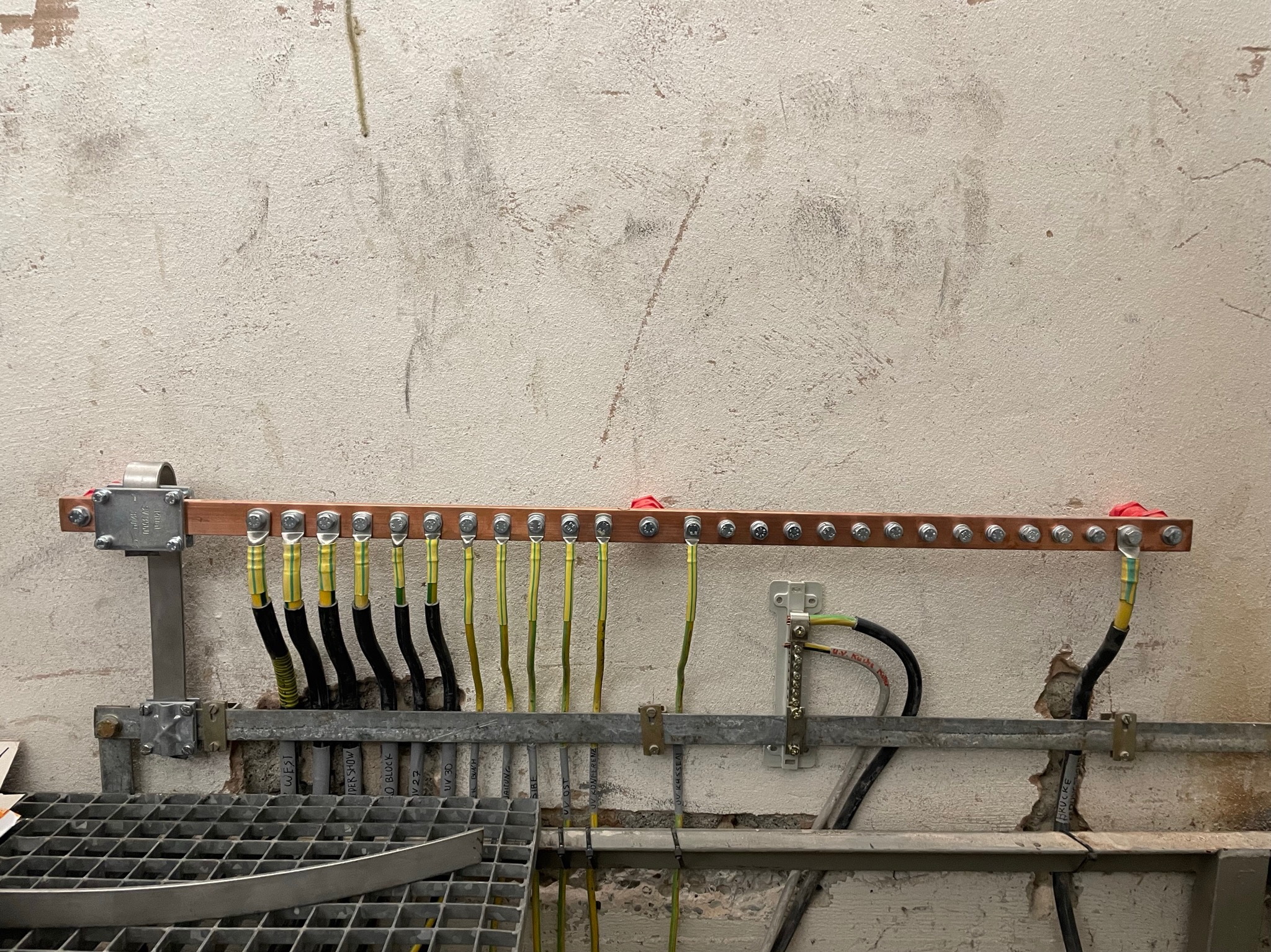 Szyna wyrównawcza potencjałów z podłączonymi przewodami ochronnymi i uziemiającymi, zamontowana na ścianie o chropowatej fakturze, widoczne metalowe elementy konstrukcyjne i fragment kraty podłogowej.