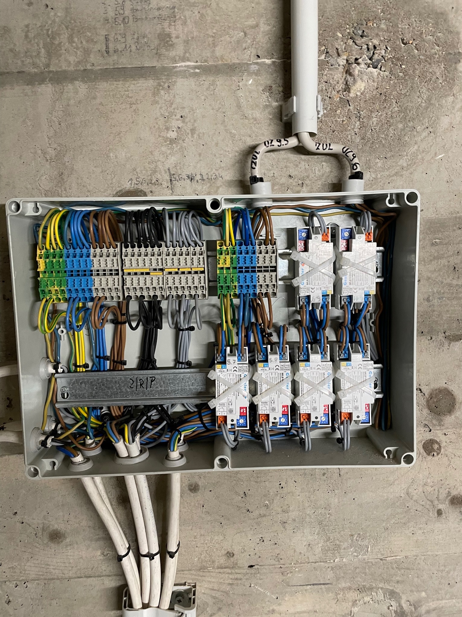 Otwarta skrzynka elektryczna z widocznymi przewodami, złączkami i zabezpieczeniami, zamontowana na surowej, betonowej ścianie z widocznymi śladami po ołówku i instalacji.