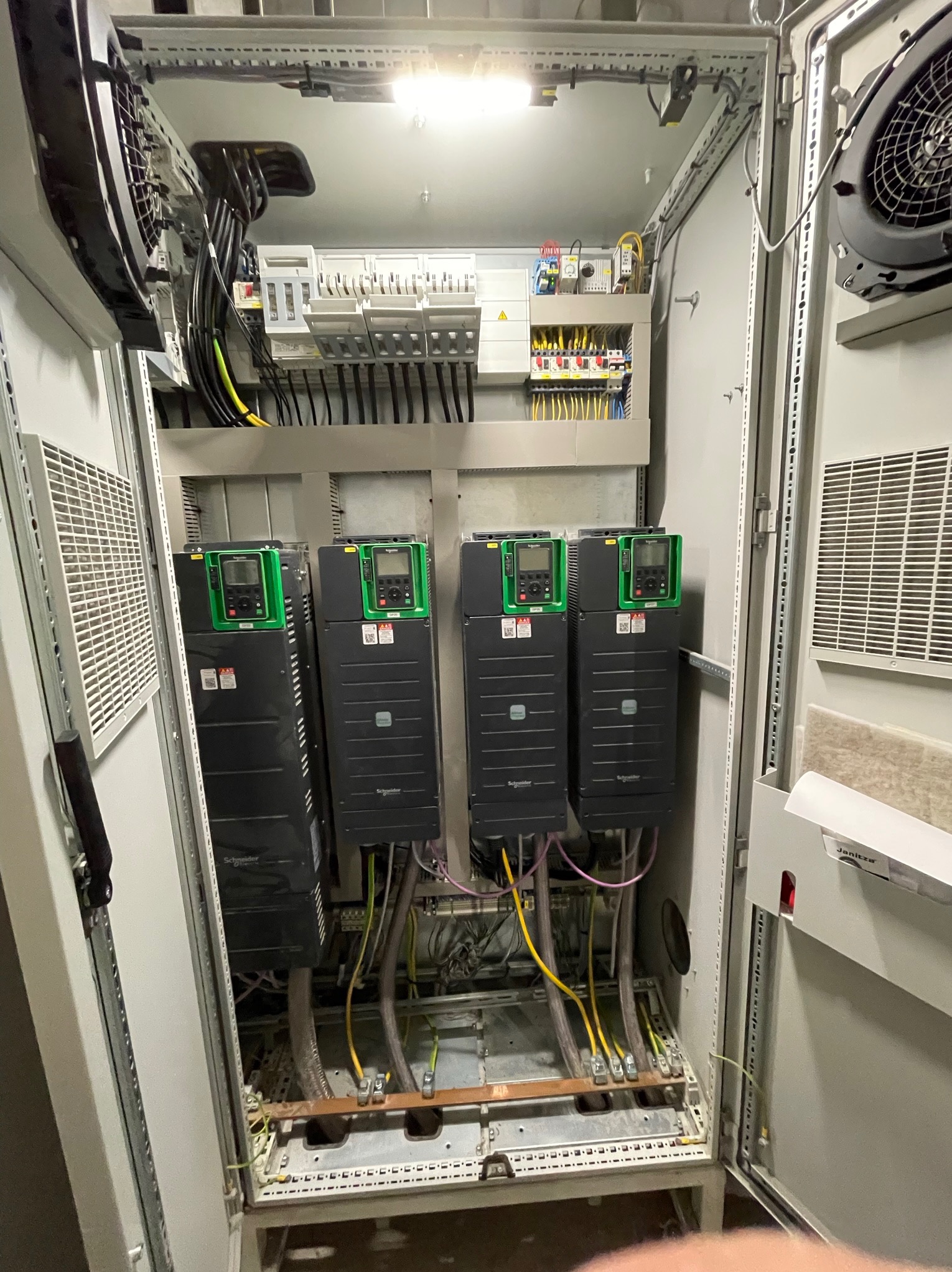 Wnętrze szafy sterowniczej z czterema falownikami Schneider Electric z zielonymi panelami, widoczne liczne przewody i elementy elektryczne, wentylatory chłodzące na górze i po bokach.
