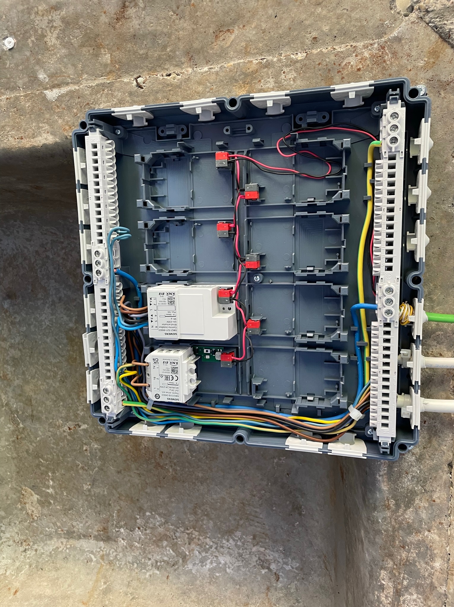 Otwarta szara skrzynka elektryczna z widocznymi przewodami, modułami sterującymi Siemens KNX E13 i listwami zaciskowymi, zamontowana na surowym, betonowym suficie.