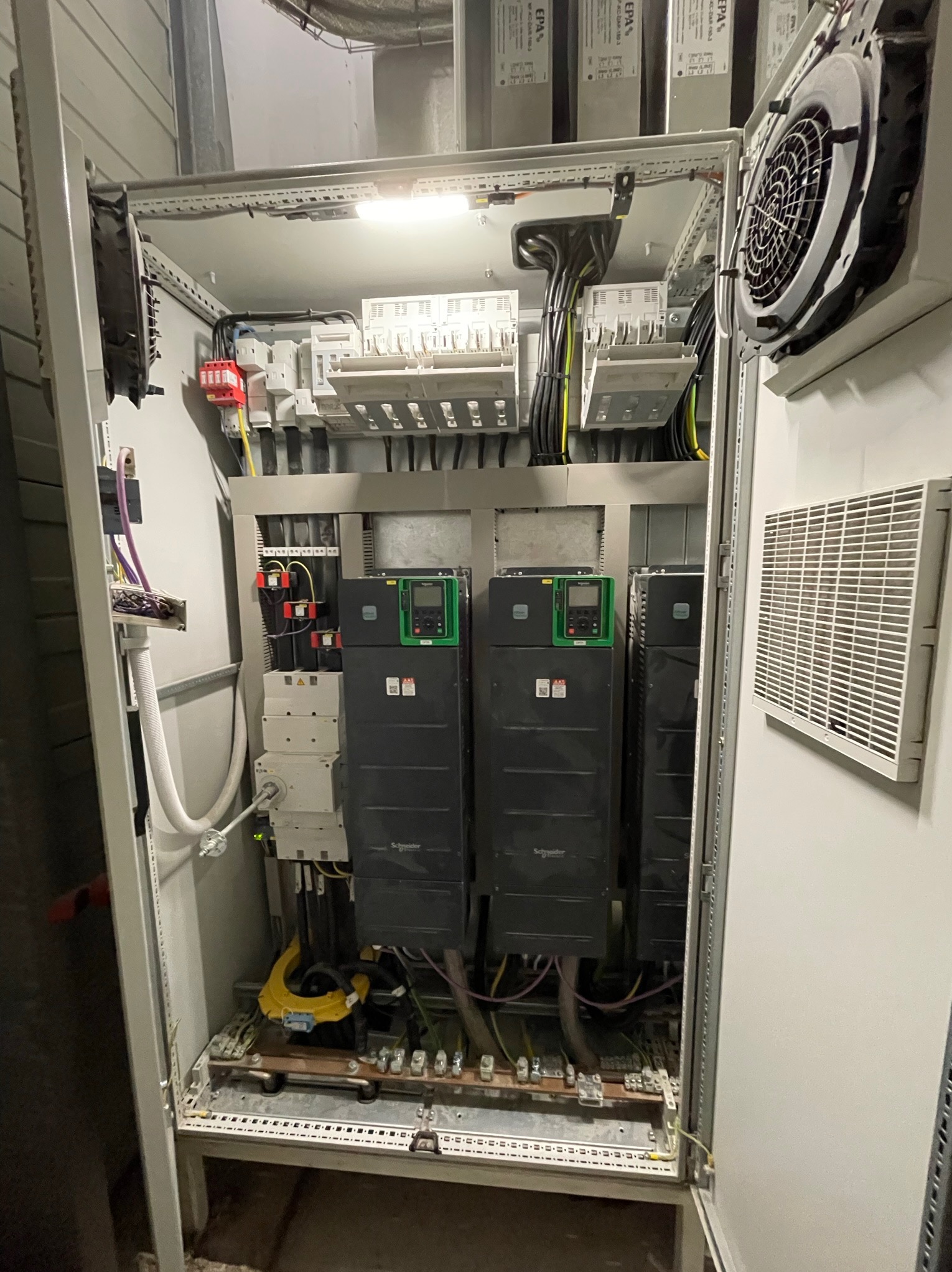 Otwarta szafa sterownicza z zainstalowanymi sterownikami silników Schneider Electric, widoczne kable, bezpieczniki i wentylator chłodzący na drzwiach szafy.