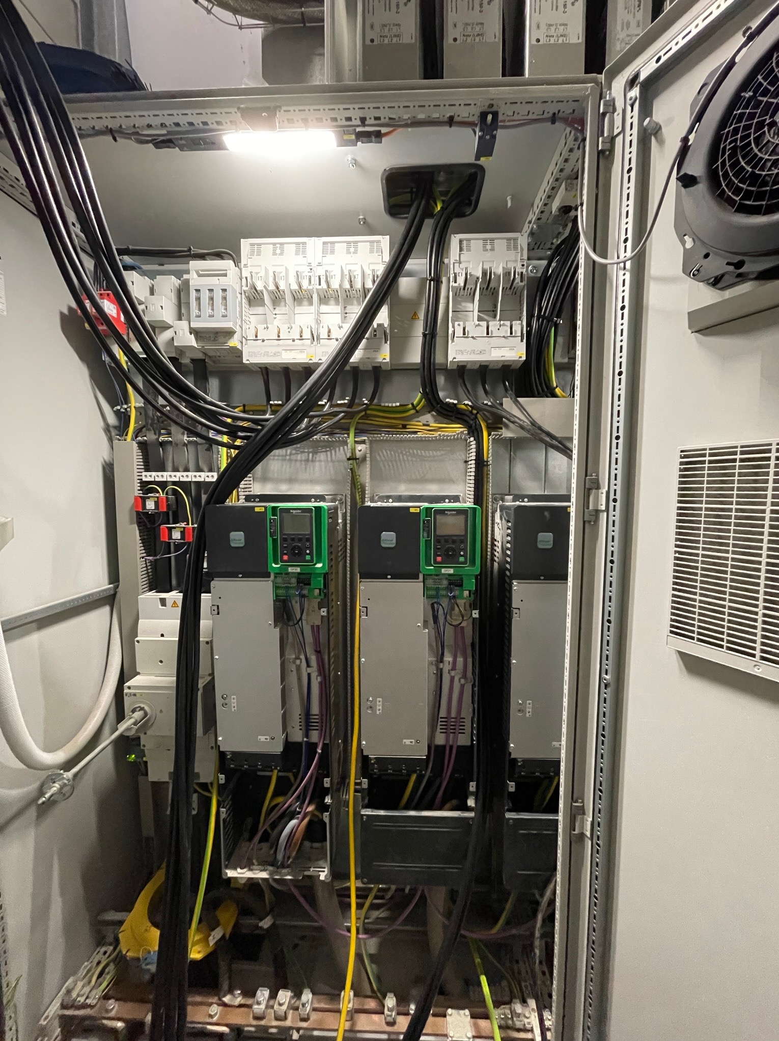 Otwarta szafa elektryczna z widocznymi bezpiecznikami, przewodami, sterownikami silników w obudowach i wentylatorem chłodzącym, oświetlona od góry.