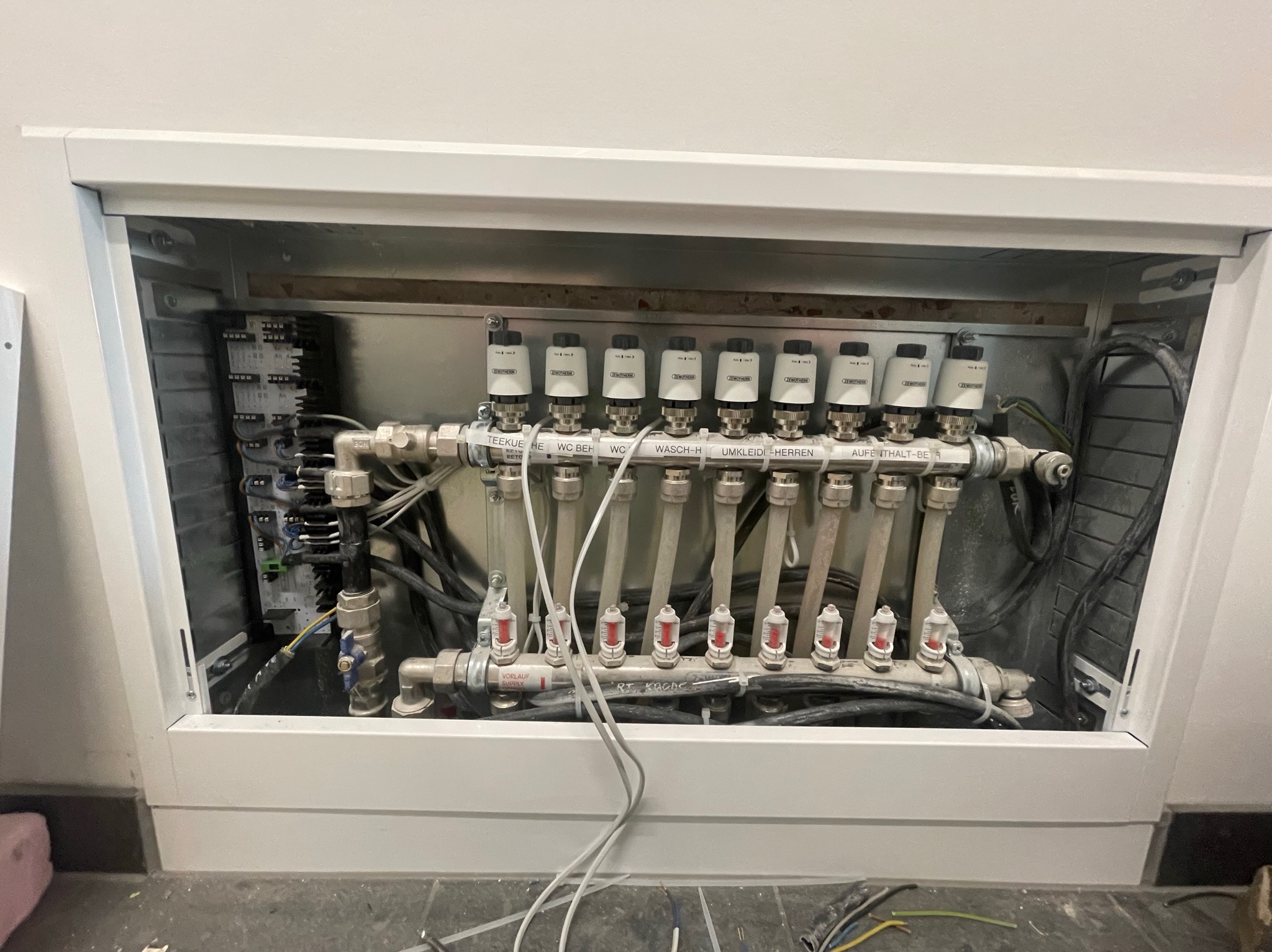 Rozdzielacz ogrzewania podłogowego w białej szafce z widocznymi zaworami termostatycznymi i przewodami elektrycznymi, zainstalowany przy podłodze.