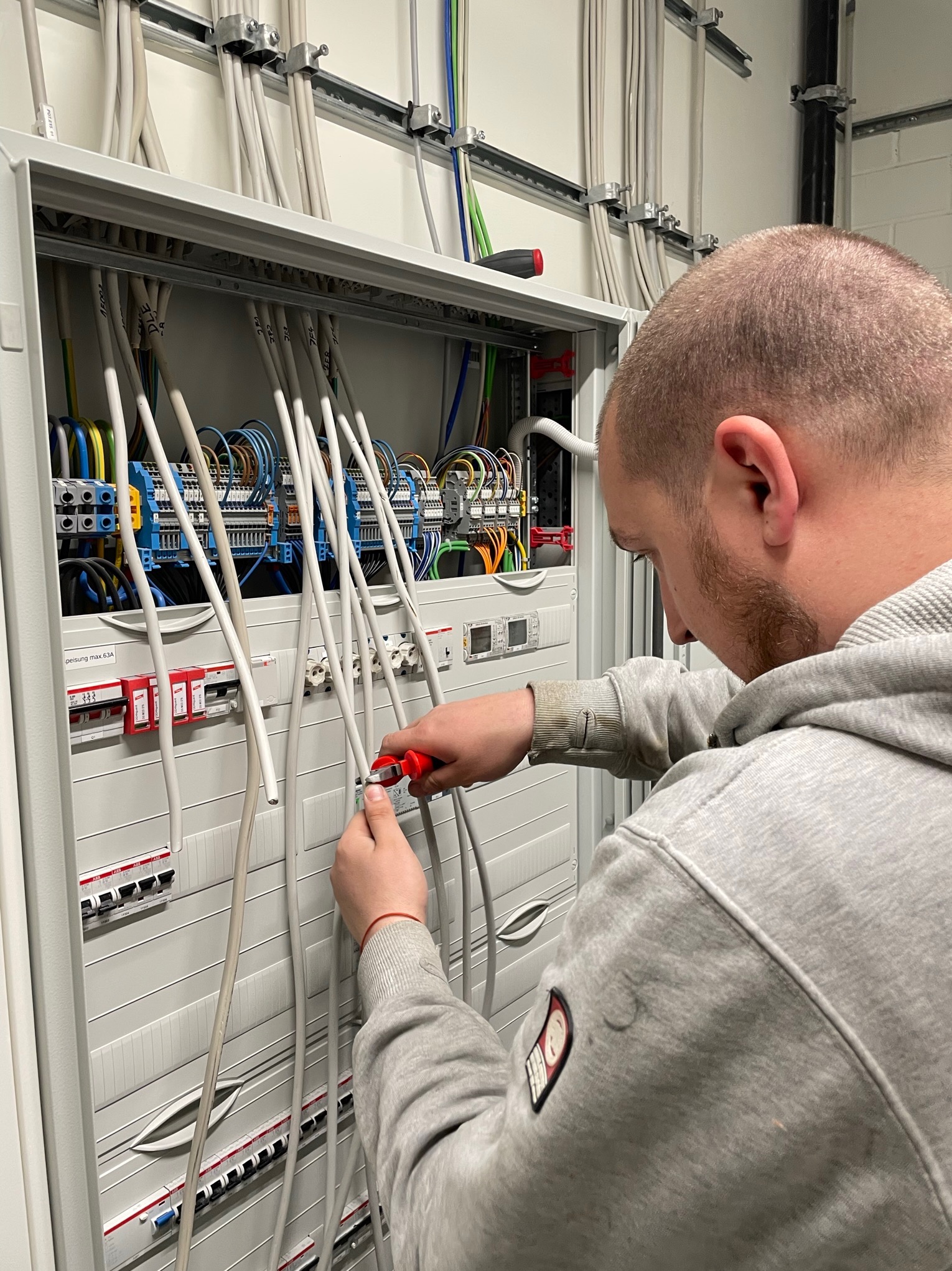 Elektryk w szarej bluzie obcina przewód w rozdzielnicy elektrycznej za pomocą czerwonych obcinaczek, widoczne liczne kable i zabezpieczenia.