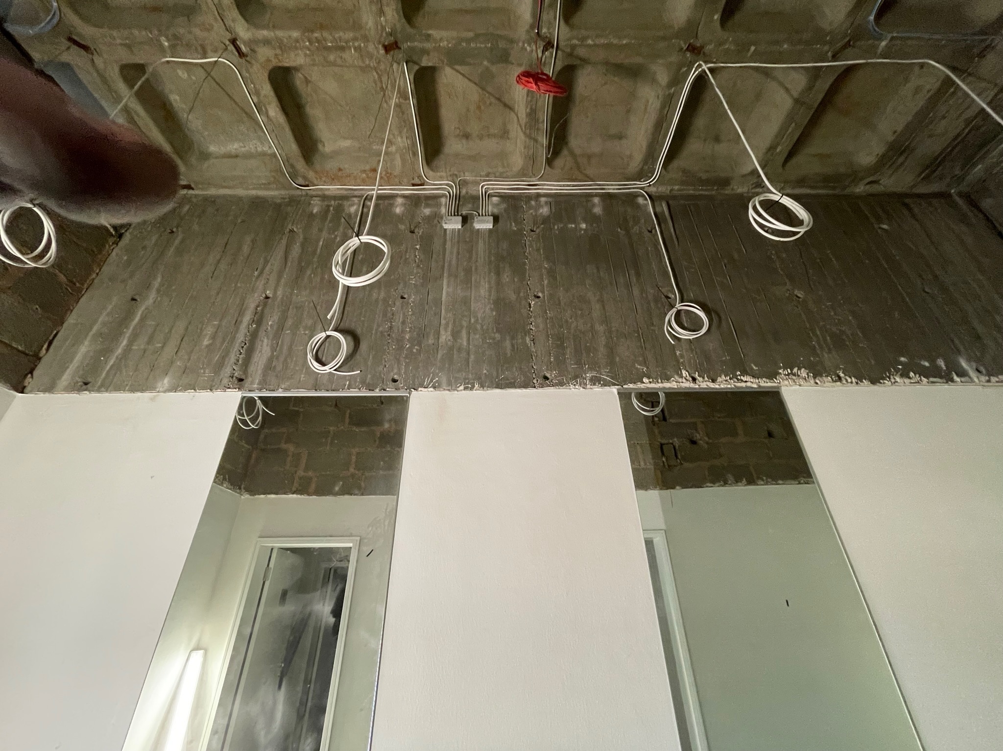 Betonowy sufit z widocznymi przewodami elektrycznymi, puszkami instalacyjnymi i fragmentami ścian z lustrami, w trakcie prac wykończeniowych.