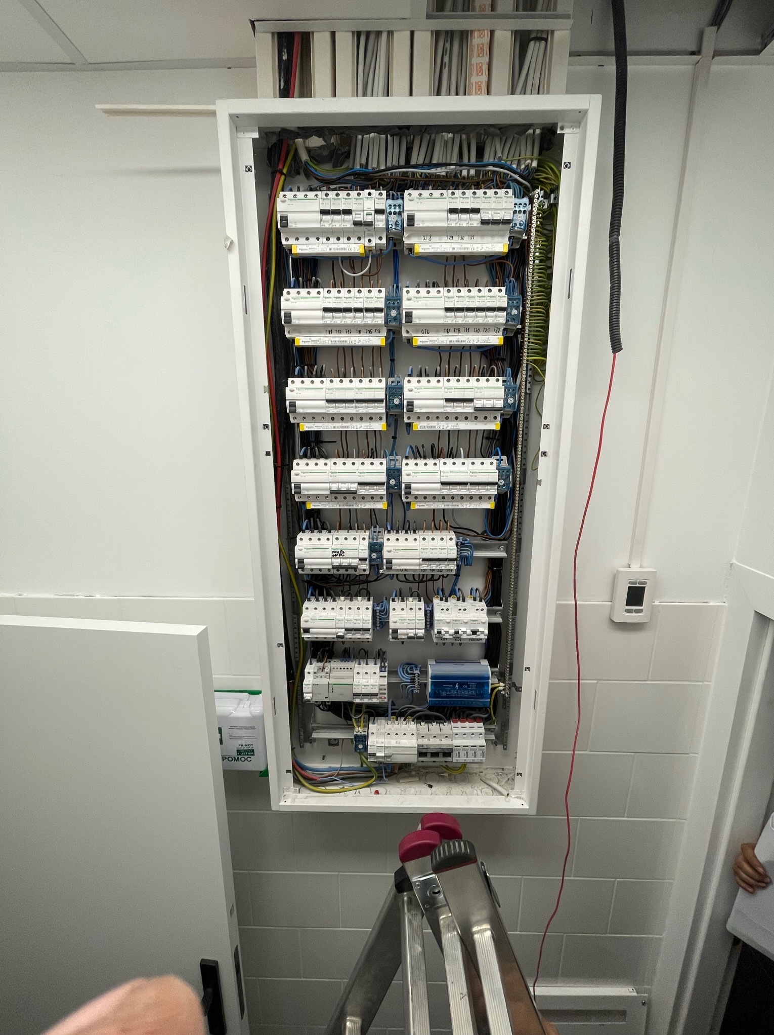 Otwarta skrzynka rozdzielcza z bezpiecznikami i przewodami w jasnym pomieszczeniu, widoczna drabina i fragment dłoni w dolnej części kadru.