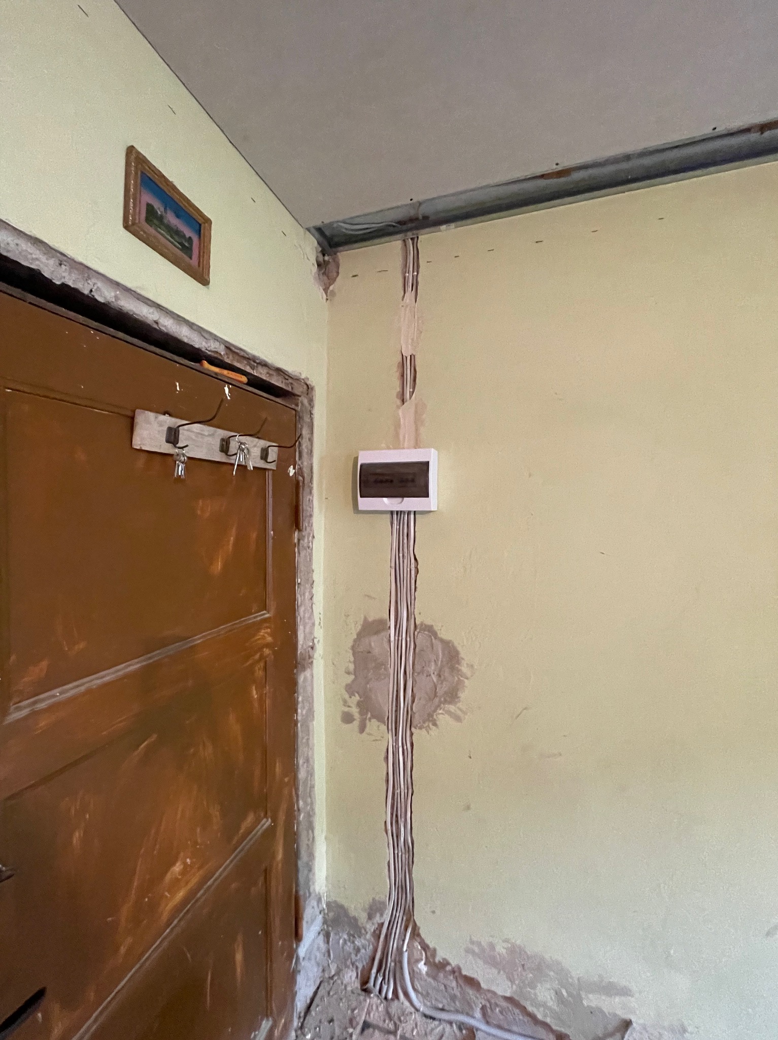 Montaż podtynkowy skrzynki elektrycznej z przewodami w bruździe ściany w trakcie remontu, widoczna stara rama drzwiowa i wystające przewody.