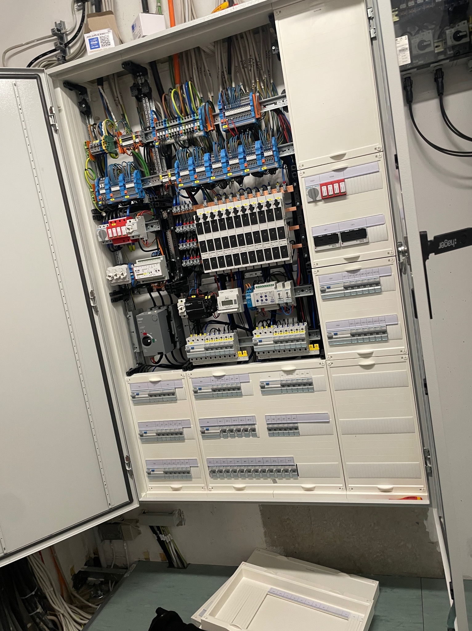 Otwarta szafa elektryczna z licznymi przewodami, bezpiecznikami i przełącznikami, widoczne także elementy montażowe i oznaczenia, na podłodze leży biała skrzynka