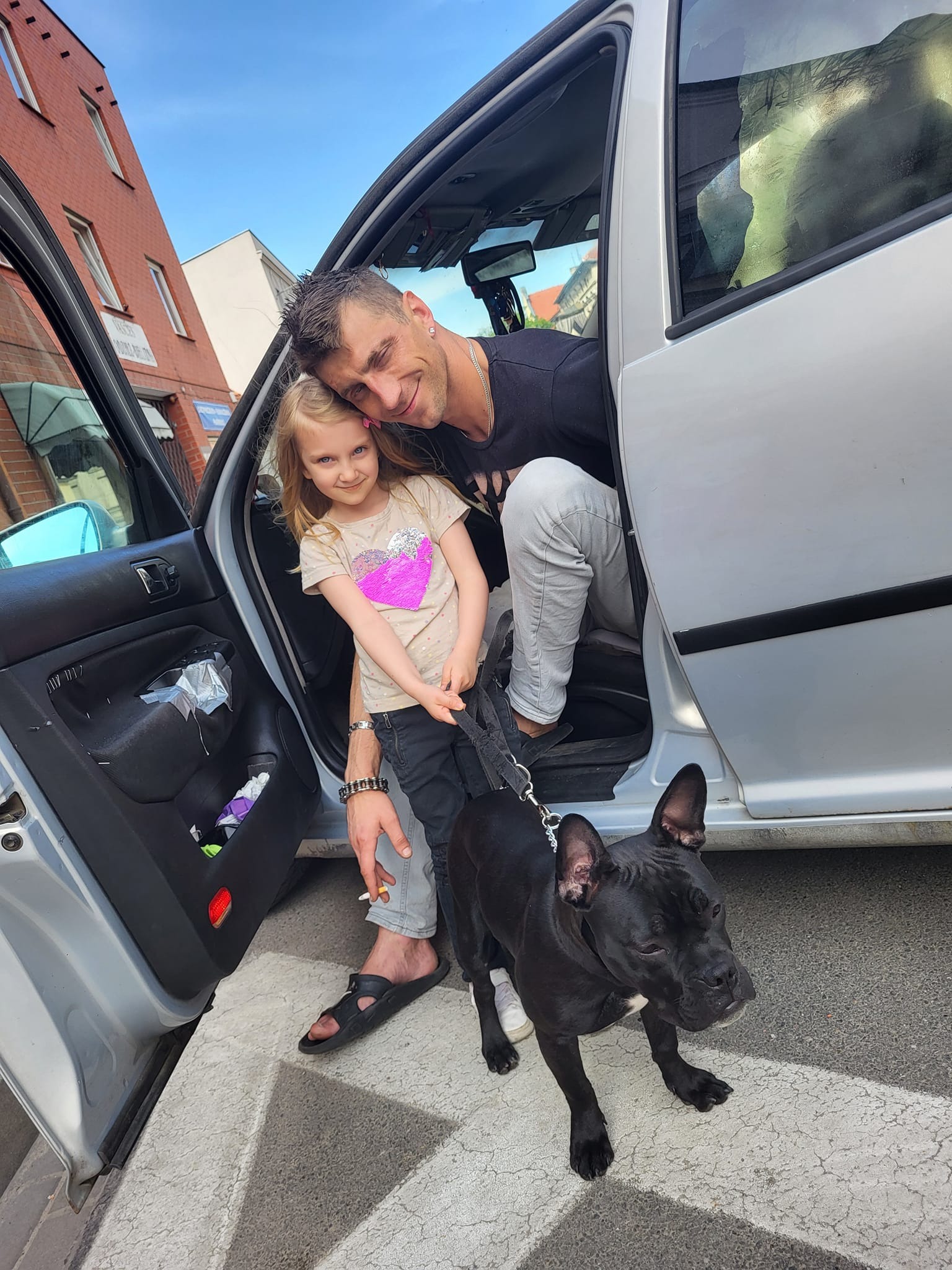 Uśmiechnięty mężczyzna z córką i psem rasy American Bully w srebrnym samochodzie z otwartymi drzwiami na tle miejskiej zabudowy.