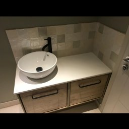 Marek Piotrowski - Nowoczesna łazienka z okrągłą umywalką nablatową, czarną baterią i szafką z drewnianymi frontami i czarnymi uchwytami, wykończona beżowymi, kwadratowymi płytkami.