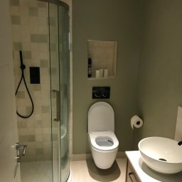 Marek Piotrowski - Nowoczesna łazienka z kabiną prysznicową typu walk-in, wiszącą toaletą z deską wolnoopadającą, umywalką nablatową i szafką pod umywalką w kolorze jasnego drewna, wykończona w stonowanej kolorystyce...