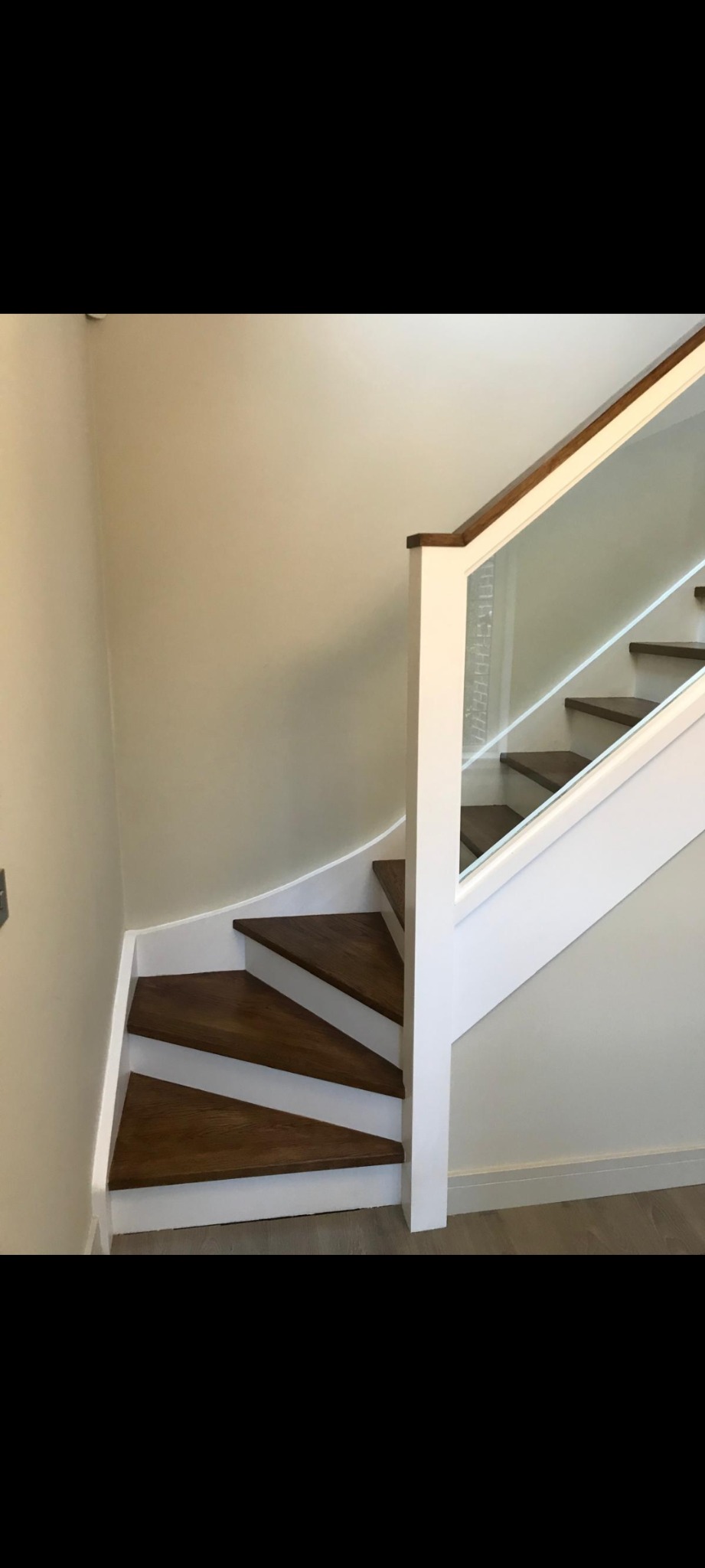 Drewniane schody z białymi podstopnicami i szklaną balustradą, zakręcające w lewo przy ścianie o jasnym odcieniu beżu.