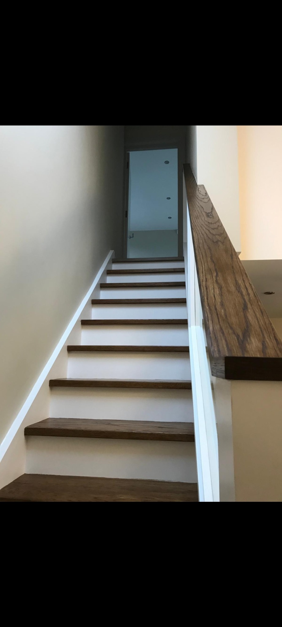 Schody z drewnianymi stopniami i białą balustradą prowadzące na wyższe piętro, widoczne trzy punkty świetlne w suficie za otwartymi drzwiami.