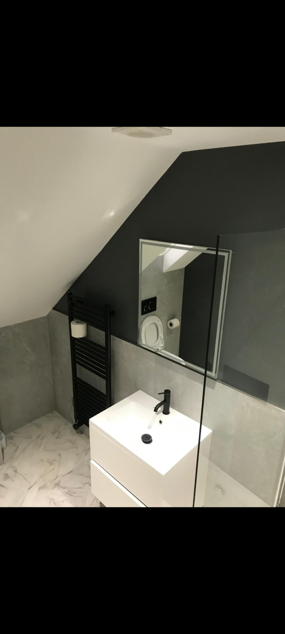 Nowoczesna łazienka na poddaszu z białą umywalką na szafce, czarną armaturą, grzejnikiem drabinkowym i lustrem z podświetleniem, odbijającym toaletę; marmurowe płytki na podłodze i ścianach.