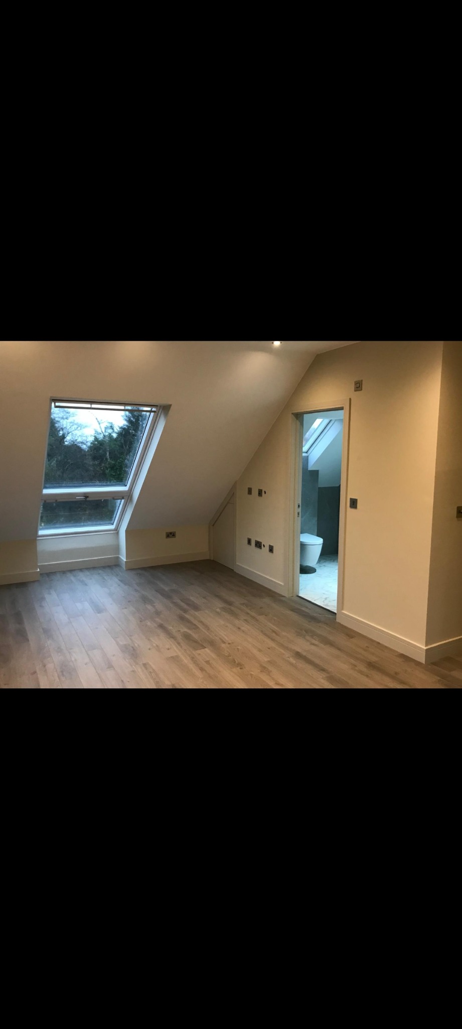Poddasze po remoncie z oknem dachowym, panelami podłogowymi w odcieniach szarości i wejściem do wyremontowanej łazienki z toaletą.