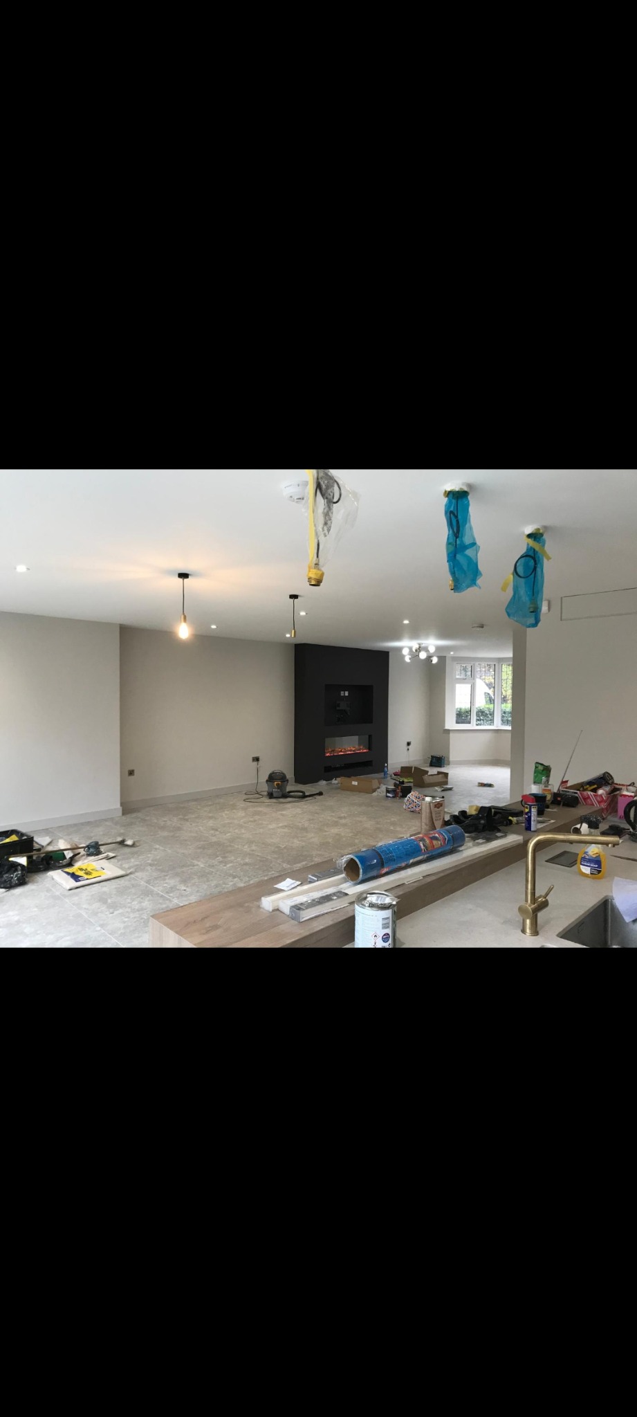 Wnętrze w trakcie remontu: szare ściany, częściowo położona podłoga z płytek, zabudowa kominkowa w kolorze antracytowym, widoczne narzędzia i materiały budowlane, zabezpieczone lampy sufitowe folią...