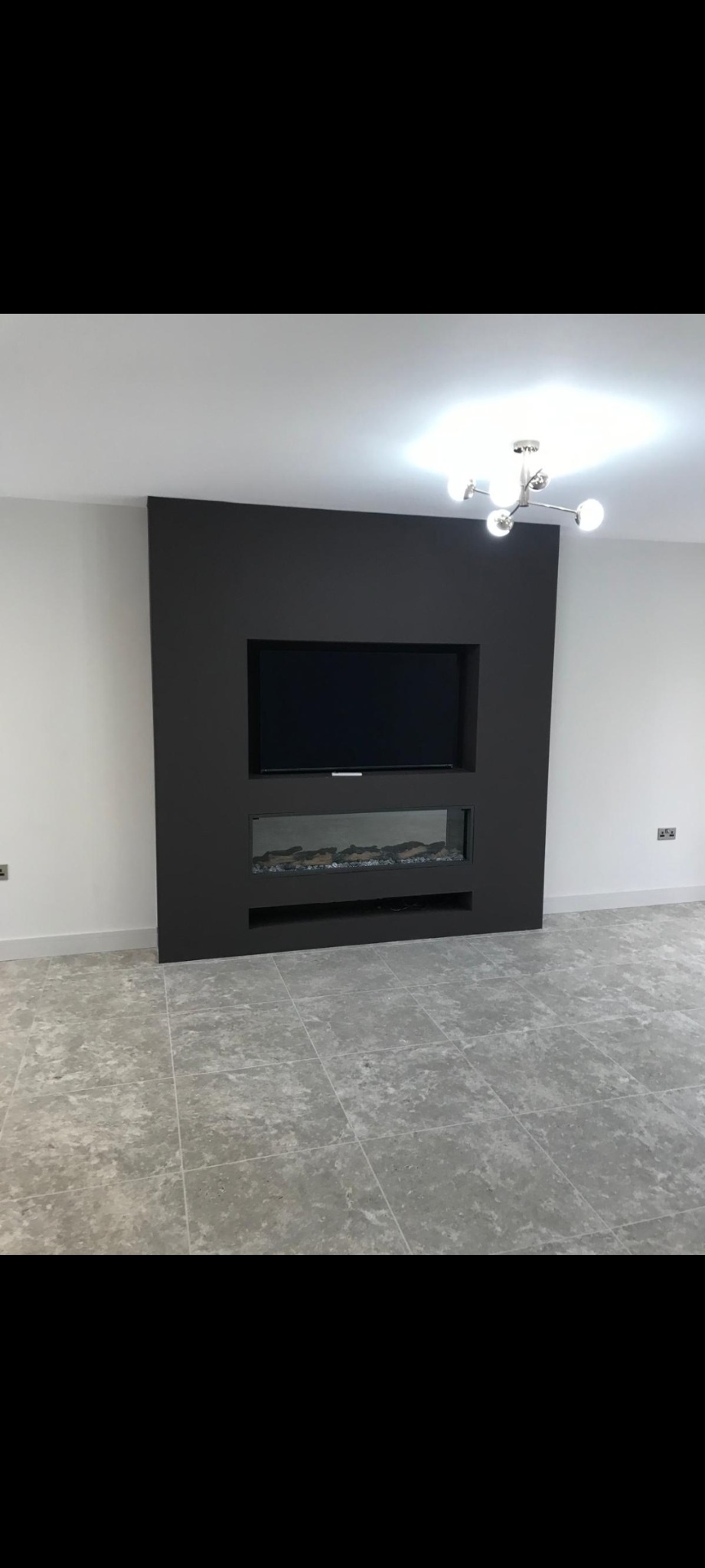 Minimalistyczna aranżacja ściany z wnęką na telewizor i elektryczny kominek, szara zabudowa na białym tle ścian, szara podłoga z płytek, nowoczesna lampa sufitowa.