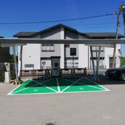 Modern Dream - Zadaszone stanowisko ładowania pojazdów elektrycznych z wyznaczonymi miejscami parkingowymi oznaczonymi na zielono, na tle budynku z parkingiem i słupem energetycznym.