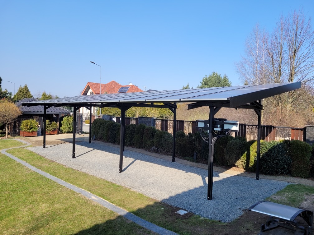 Zadaszenie na samochody z panelami słonecznymi w ogrodzie, widoczne skrzynki z instalacją elektryczną, w tle dom z panelami fotowoltaicznymi na dachu.