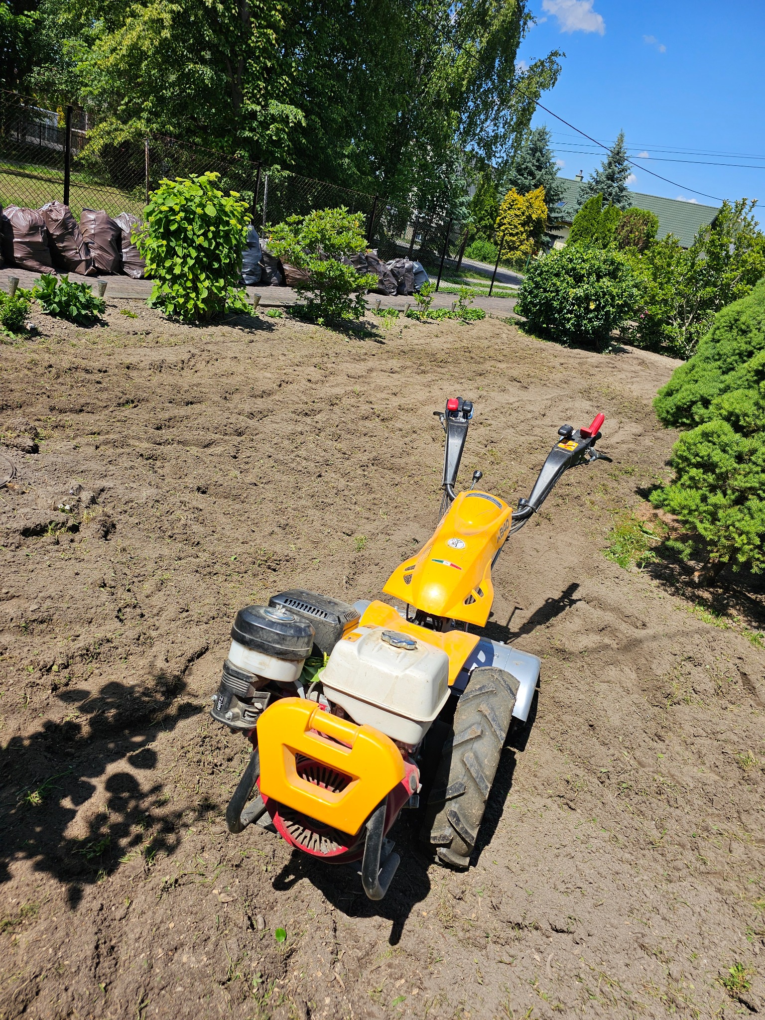 Żółto-czarny glebogryzarek spalinowy na przygotowanym podłożu z widocznymi workami z ziemią w tle, sugerujący przygotowanie terenu pod trawnik lub ogród.