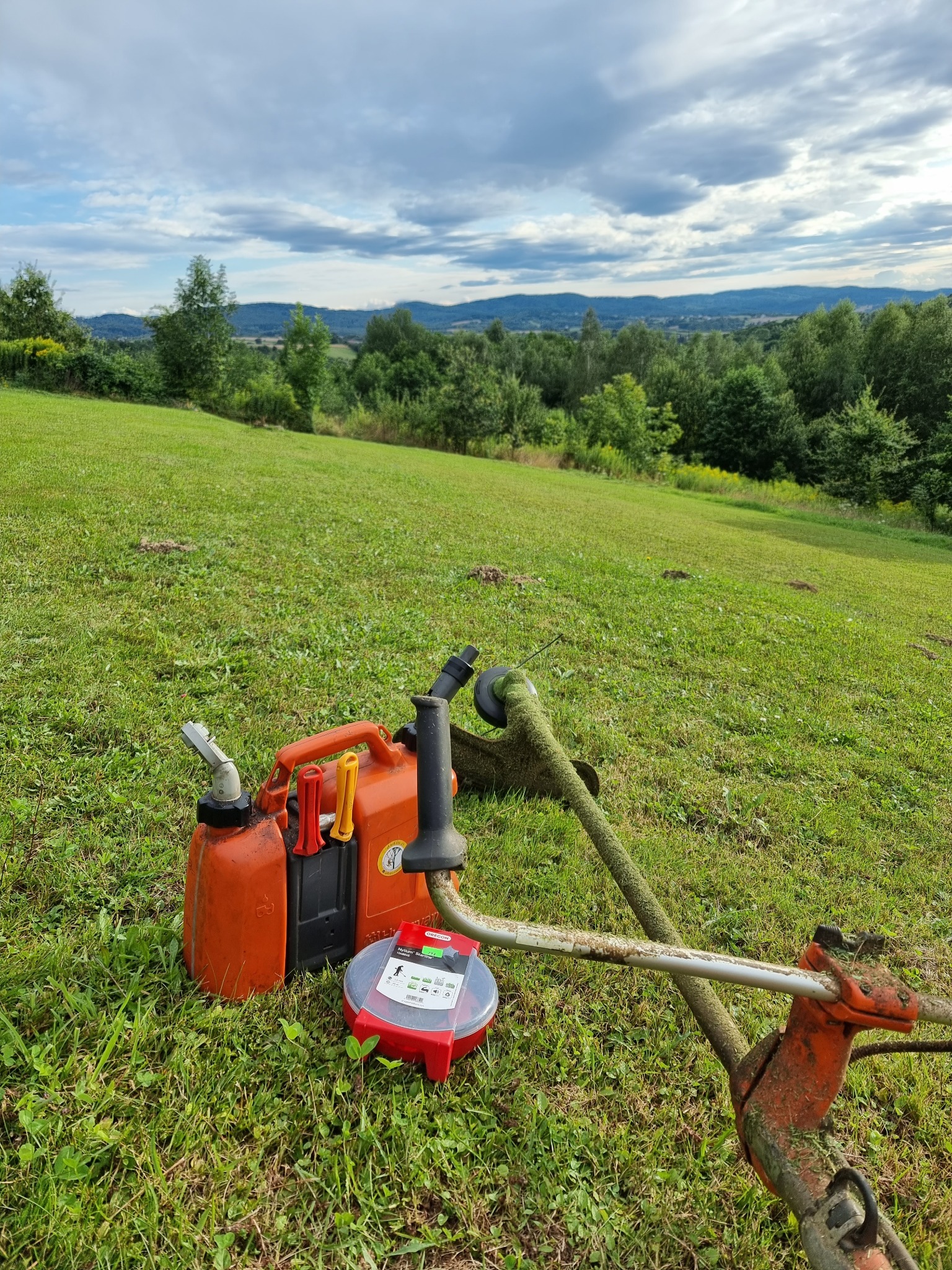 Pomarańczowy kanister z paliwem i akcesoria do podkaszarki spalinowej leżą na trawie. W tle widoczny rozległy, zielony teren i zalesione wzgórza pod pochmurnym niebem.