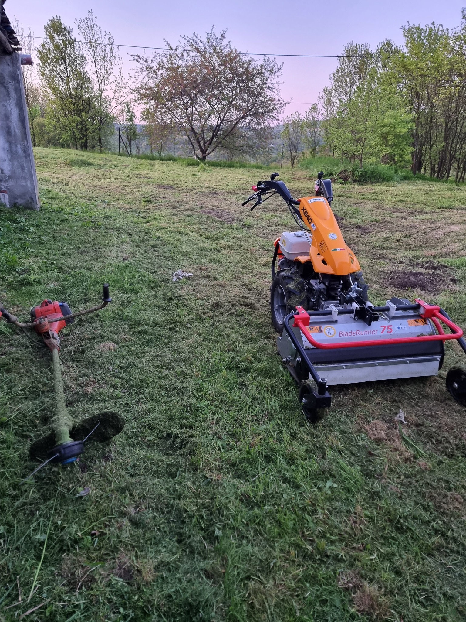 Pomarańczowa kosiarka bijakowa BladeRunner 75 i podkaszarka spalinowa na trawniku po koszeniu, widoczne ślady cięcia trawy.