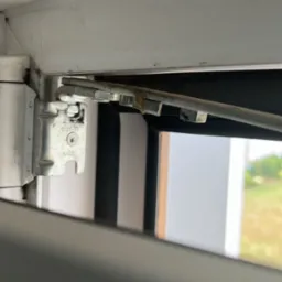 Szczegółowe ujęcie mechanizmu otwierania okna PCV, widoczne zabrudzenia i ślady użytkowania na elementach metalowych, zarys krajobrazu za oknem.