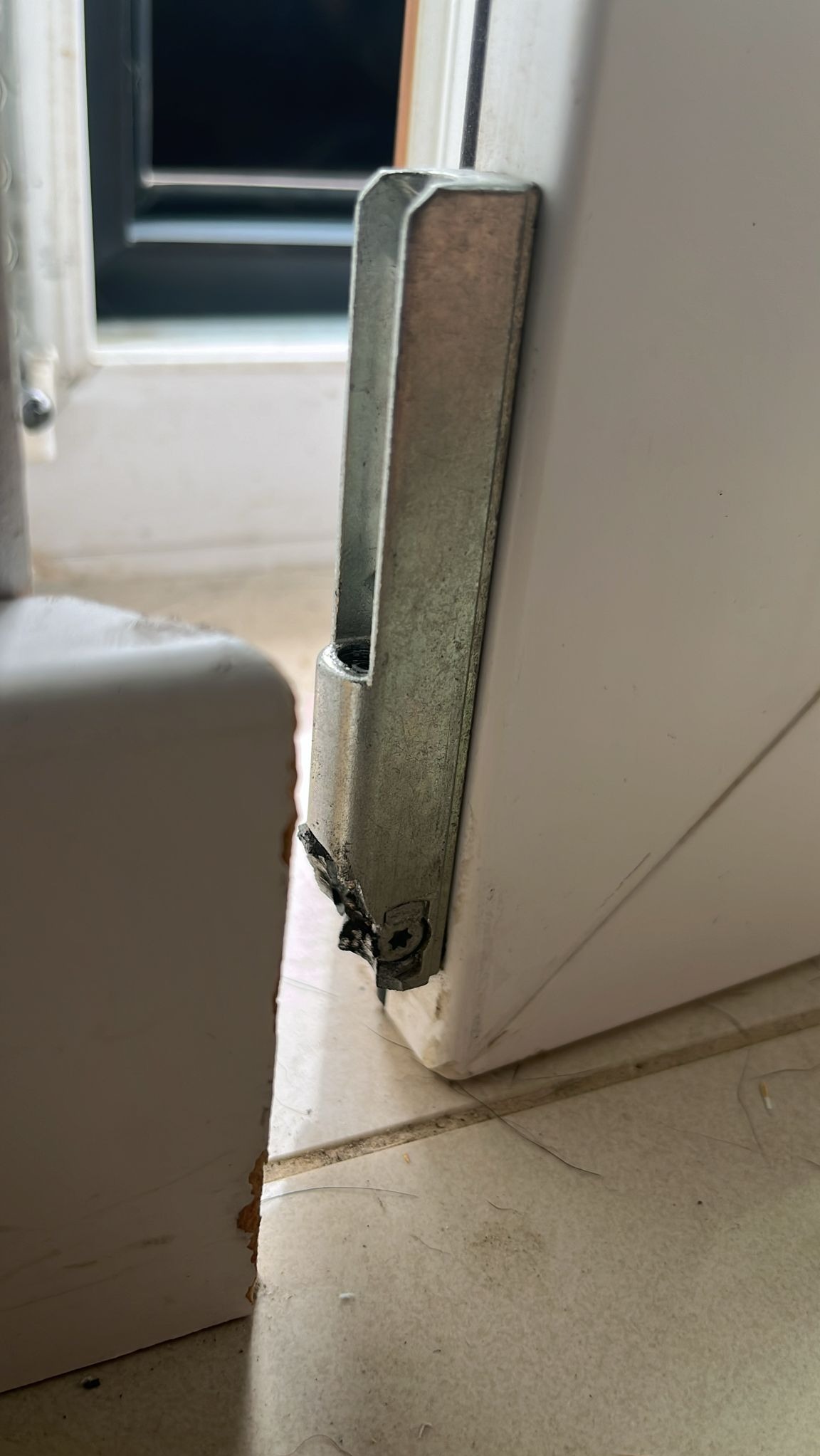 Uszkodzone okucie okienne na białym oknie PCV, widoczne ślady uszkodzeń i zużycia, brudna podłoga w tle.