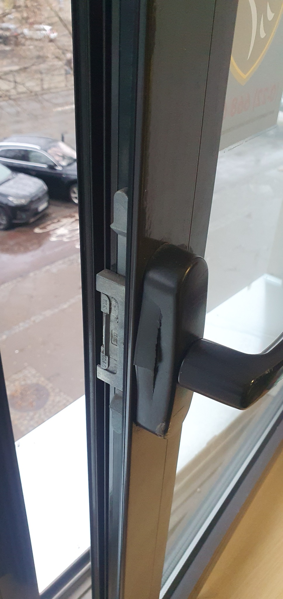 Czarny profil okienny z widocznym mechanizmem blokującym i uszkodzoną klamką, na zewnątrz widoczne zaparkowane samochody.