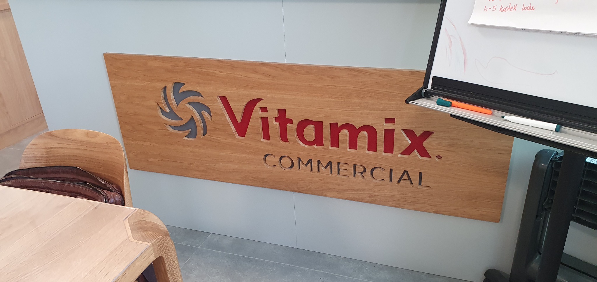 Drewniana tablica z wyciętym logo i napisem Vitamix Commercial, umieszczona na jasnej ścianie, obok stoi krzesło i tablica suchościeralna z markerami.