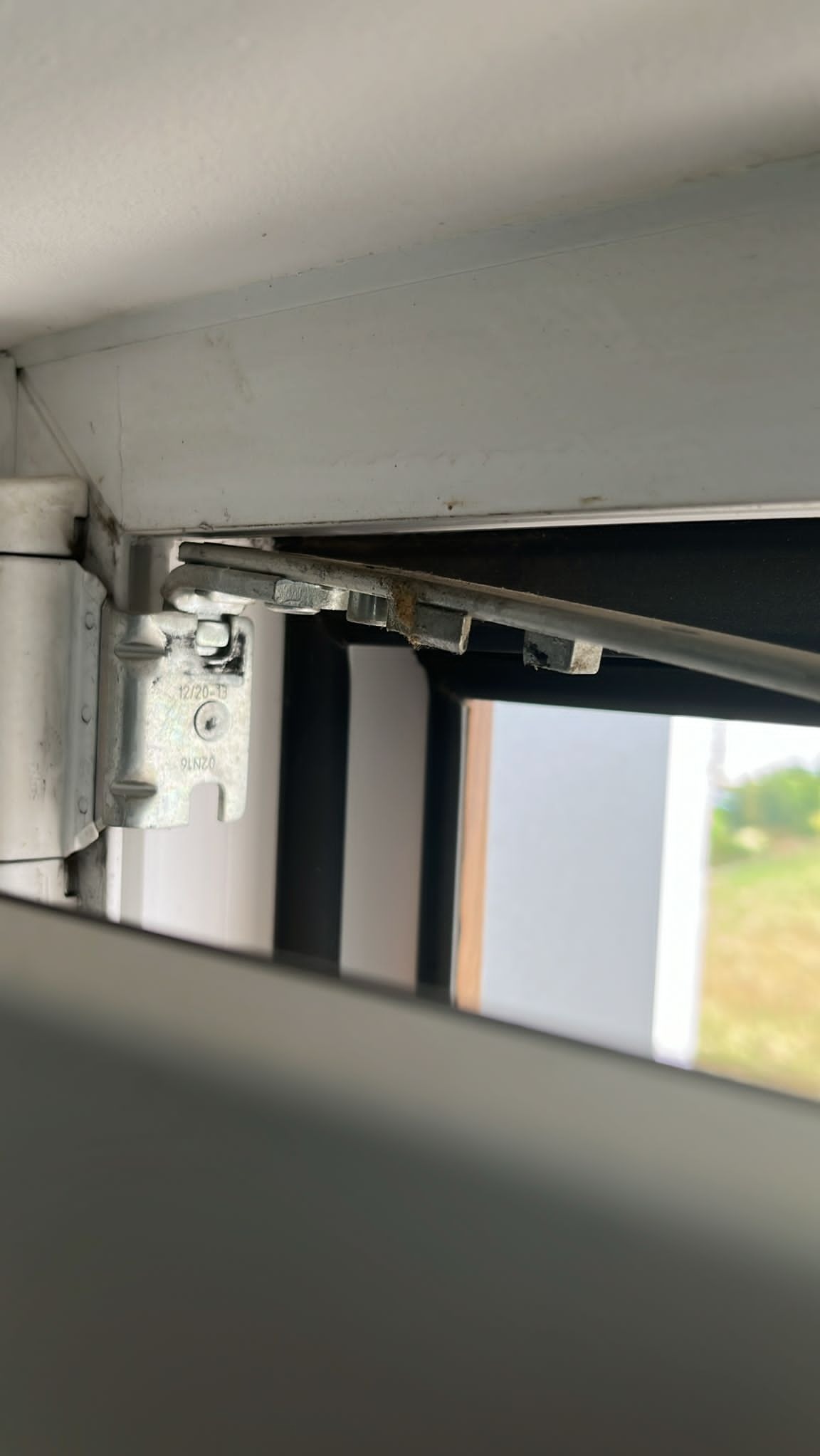 Szczegółowe ujęcie mechanizmu otwierania okna PCV, widoczne zabrudzenia i ślady użytkowania na elementach metalowych, zarys krajobrazu za oknem.