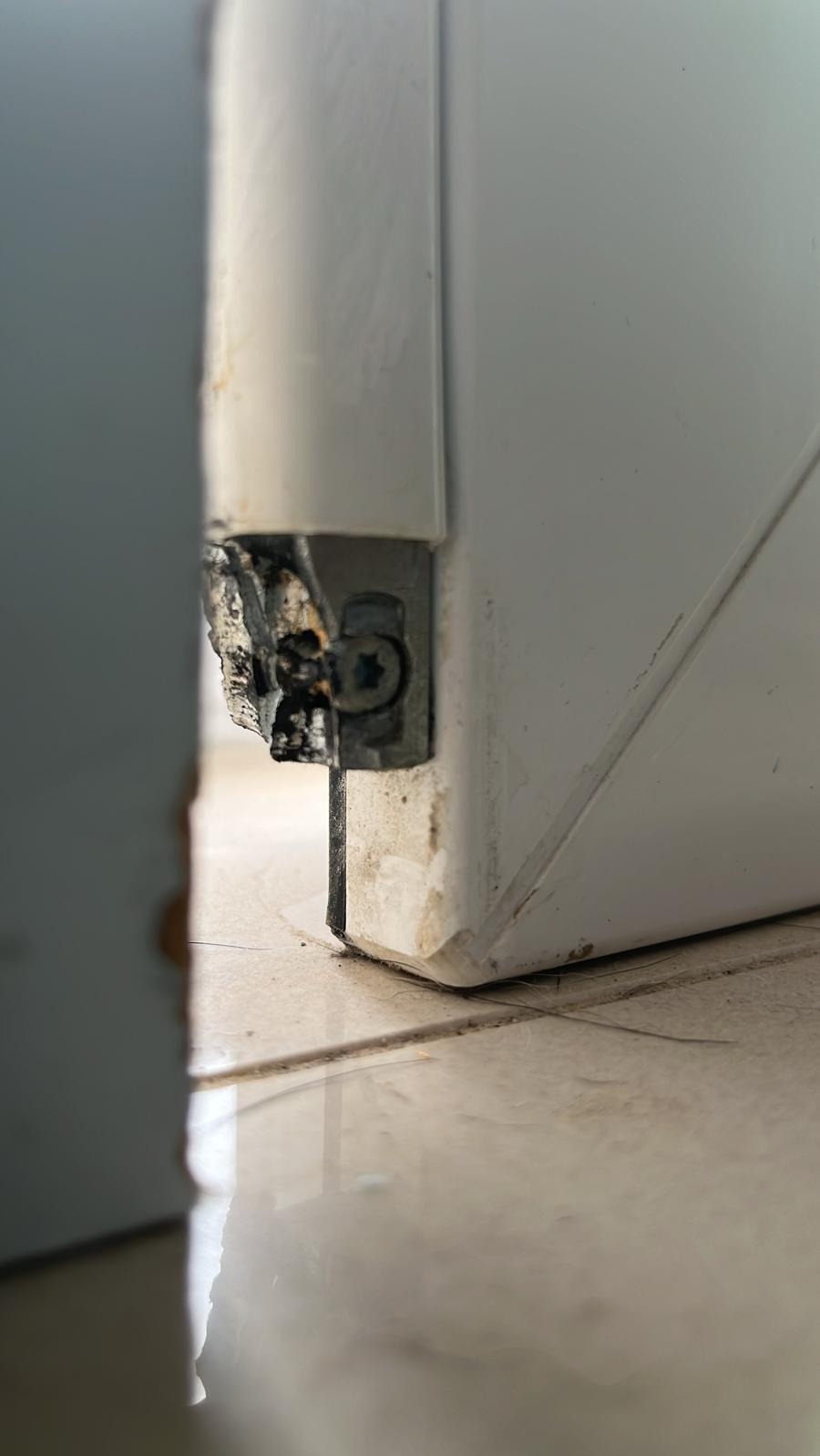 Uszkodzony narożnik białej ramy okiennej z widocznym śrubą mocującą, ubytki materiału i zabrudzenia na styku z podłogą wyłożoną jasnymi płytkami.