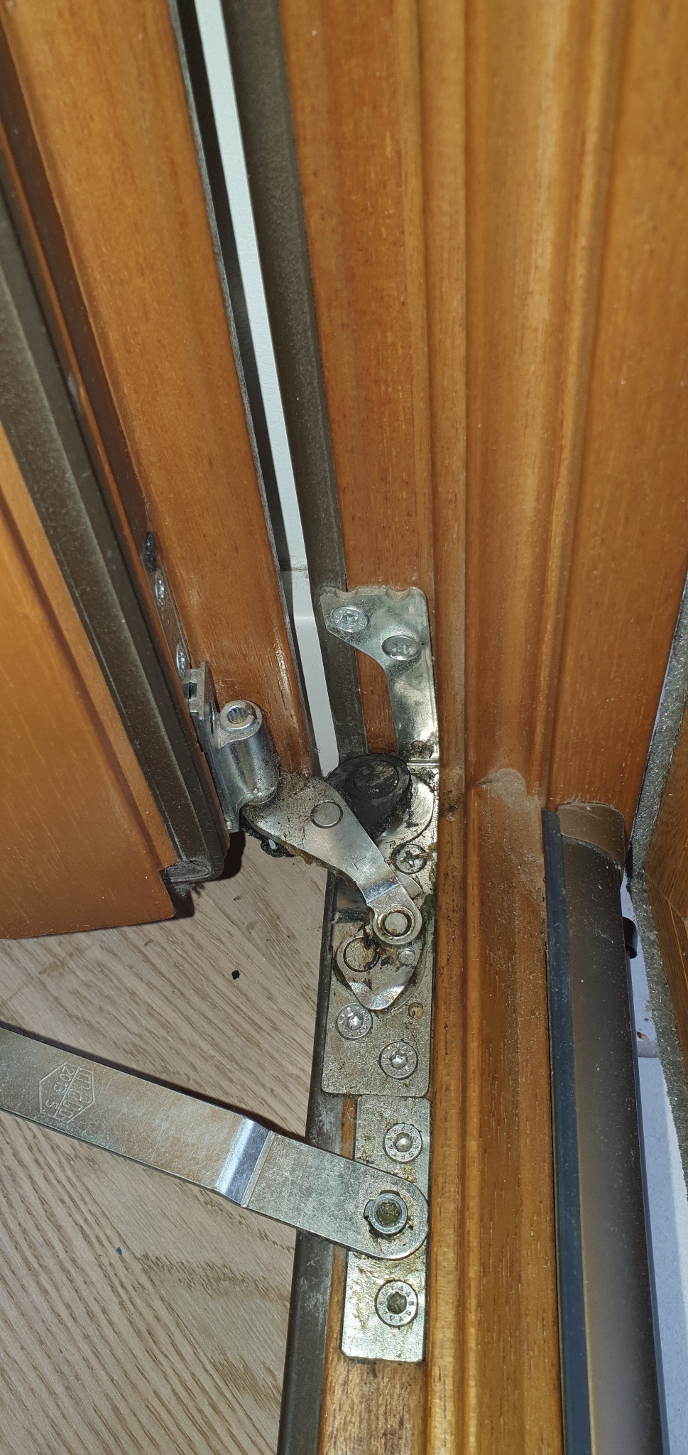 Szczegółowe ujęcie mechanizmu okna drewnianego z widocznymi śrubami i elementami metalowymi, sugerujące potrzebę regulacji lub naprawy.