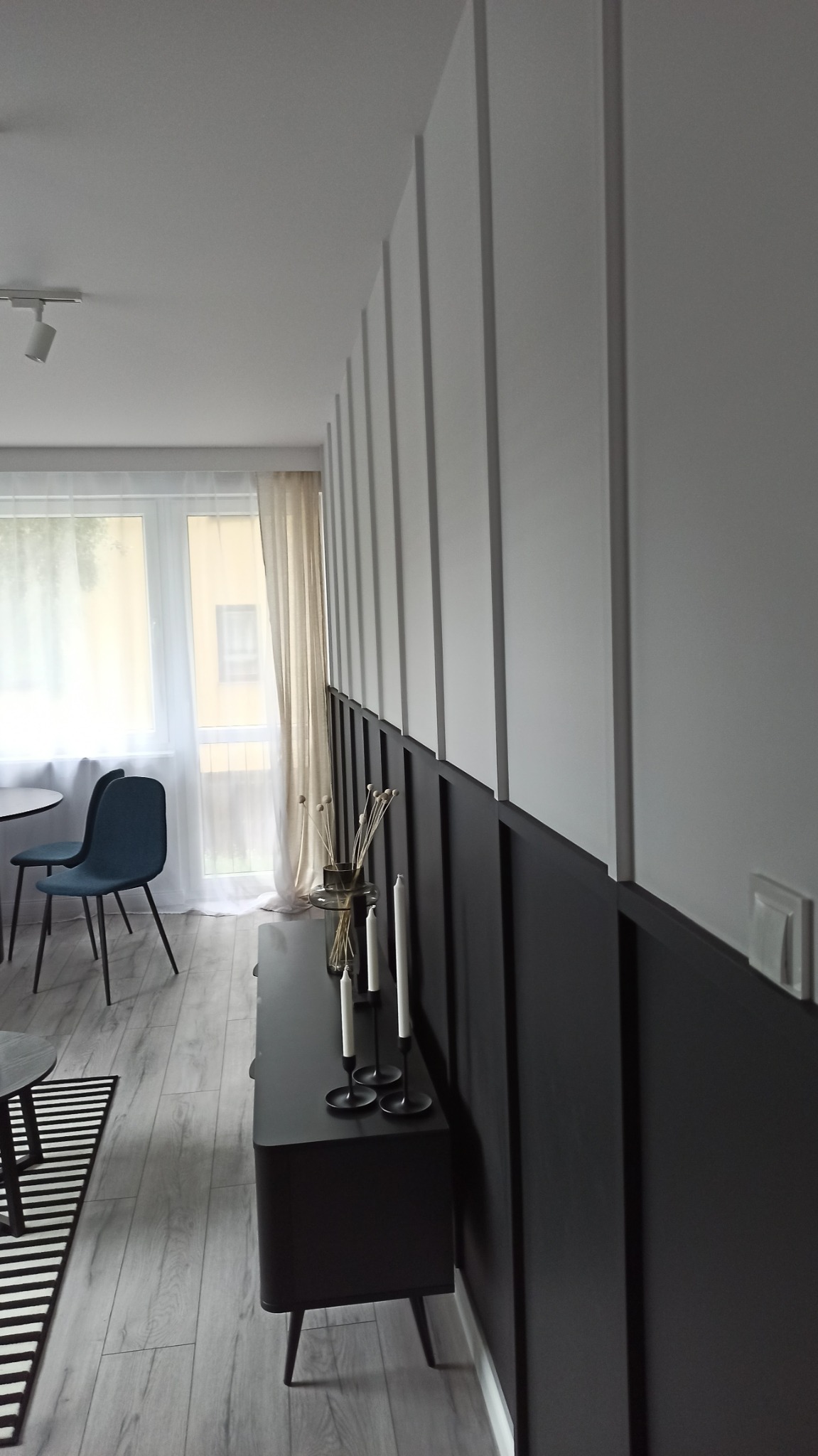 Nowoczesne szare panele ścienne z czarnym pasem dolnym i białymi pionowymi listwami, minimalistyczny wystrój wnętrza z czarną komodą, świecami i dekoracyjnymi gałązkami.