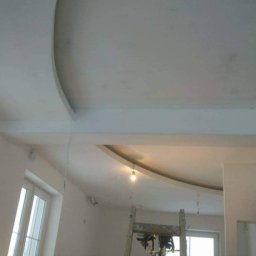 WOJT-BUD Kamil Wojtas - Wnętrze w trakcie prac wykończeniowych: widoczny sufit z elementami dekoracyjnymi z płyty karton-gips, drabina i lampa wisząca na kablu.