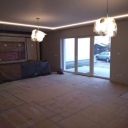 WOJT-BUD Kamil Wojtas - Pokój w trakcie prac wykończeniowych: podłoga zabezpieczona kartonem, ściany pomalowane na szaro, sufit z oświetleniem LED, wiszące lampy i widok na zewnątrz przez duże okno balkonowe.