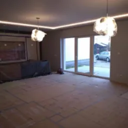 Pok&oacute;j w trakcie prac wykończeniowych: podłoga zabezpieczona kartonem, ściany pomalowane na szaro, sufit z oświetleniem LED, wiszące lampy i widok na zewnątrz przez duże okno balkonowe.