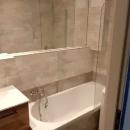 Nowoczesna łazienka z wanną obudowaną szarymi płytkami, szklanym parawanem nawannowym oraz umywalką z szafką z drewnianymi frontami i czarnymi uchwytami. Na ścianach jasnoszare płytki imitujące...