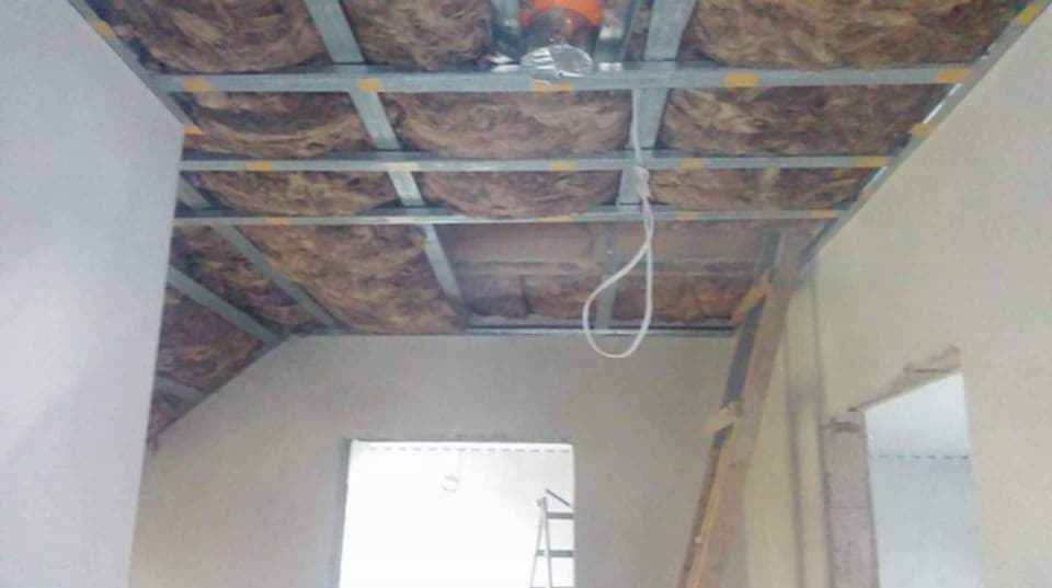 Montaż izolacji z wełny mineralnej w metalowej konstrukcji sufitu podwieszanego, widoczne fragmenty ścian i drabiny.