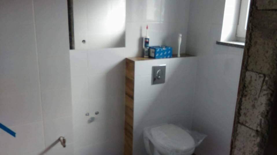 Biała łazienka w trakcie remontu: widoczne białe płytki, zabudowana spłuczka z drewnianym wykończeniem, toaleta zabezpieczona folią, oraz fragment ściany w stanie surowym.