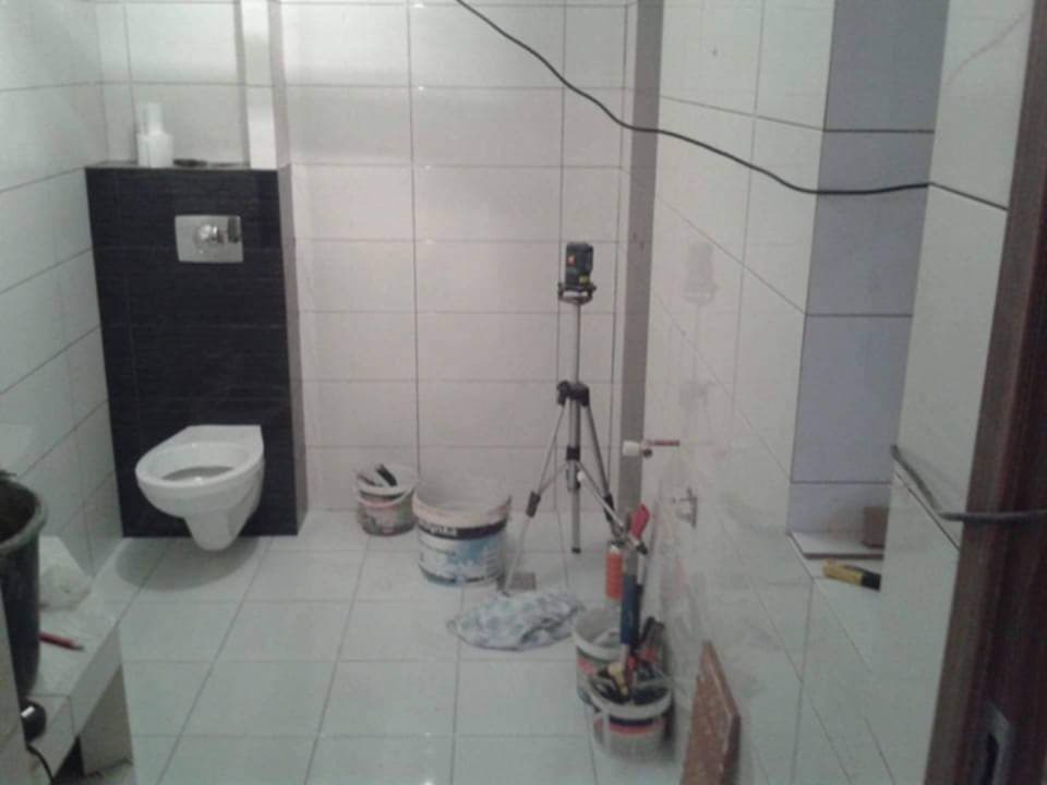 Łazienka w trakcie remontu, widoczna podwieszana toaleta na tle czarnej ściany, białe płytki na ścianach i podłodze, laser krzyżowy na statywie, wiadra z narzędziami i materiałami budowlanymi.