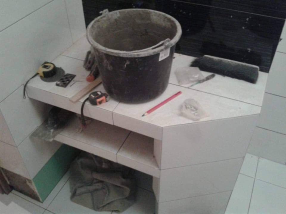 Fragment łazienki w trakcie prac: białe płytki, narożna półka z ułożonych płytek, wiadro z zaprawą, miarka, ołówek i inne narzędzia do układania płytek.