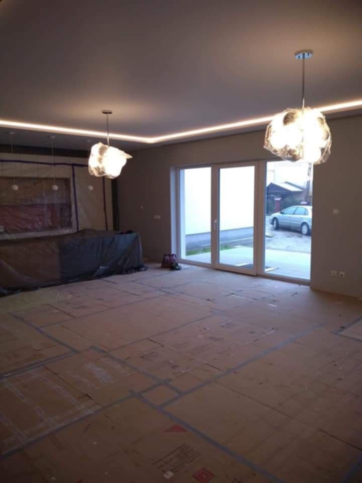 Pokój w trakcie prac wykończeniowych: podłoga zabezpieczona kartonem, ściany pomalowane na szaro, sufit z oświetleniem LED, wiszące lampy i widok na zewnątrz przez duże okno balkonowe.