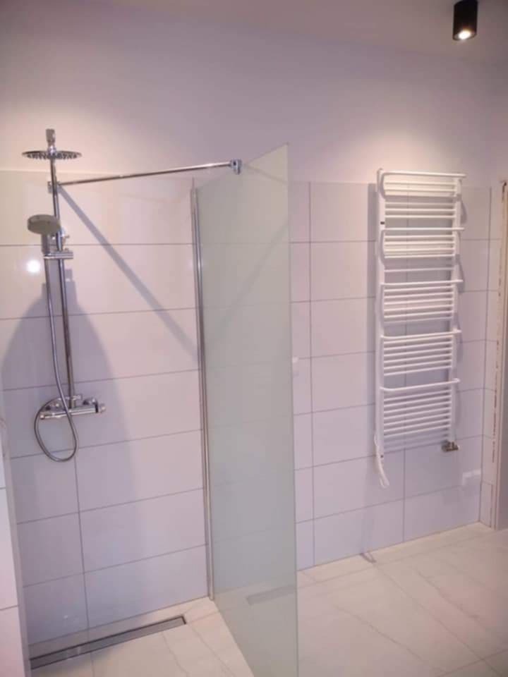 Wykończona łazienka z kabiną prysznicową typu walk-in z matową szybą, grzejnikiem drabinkowym i chromowaną armaturą na tle białych płytek.