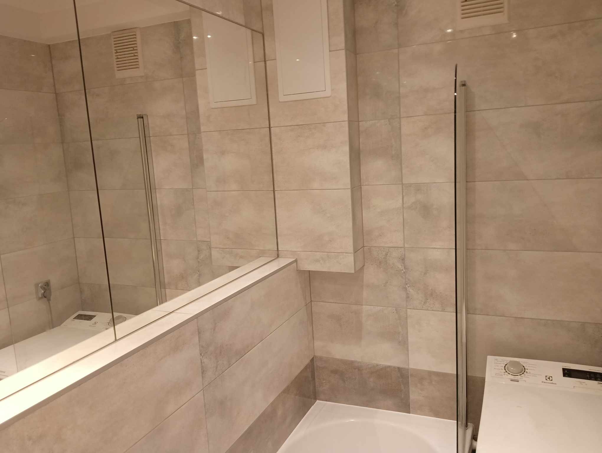 Jasna łazienka z beżowymi płytkami, kabiną prysznicową z przezroczystego szkła i pralką Electrolux w rogu, widok na wentylację i rewizje zabudowane w płytkach.