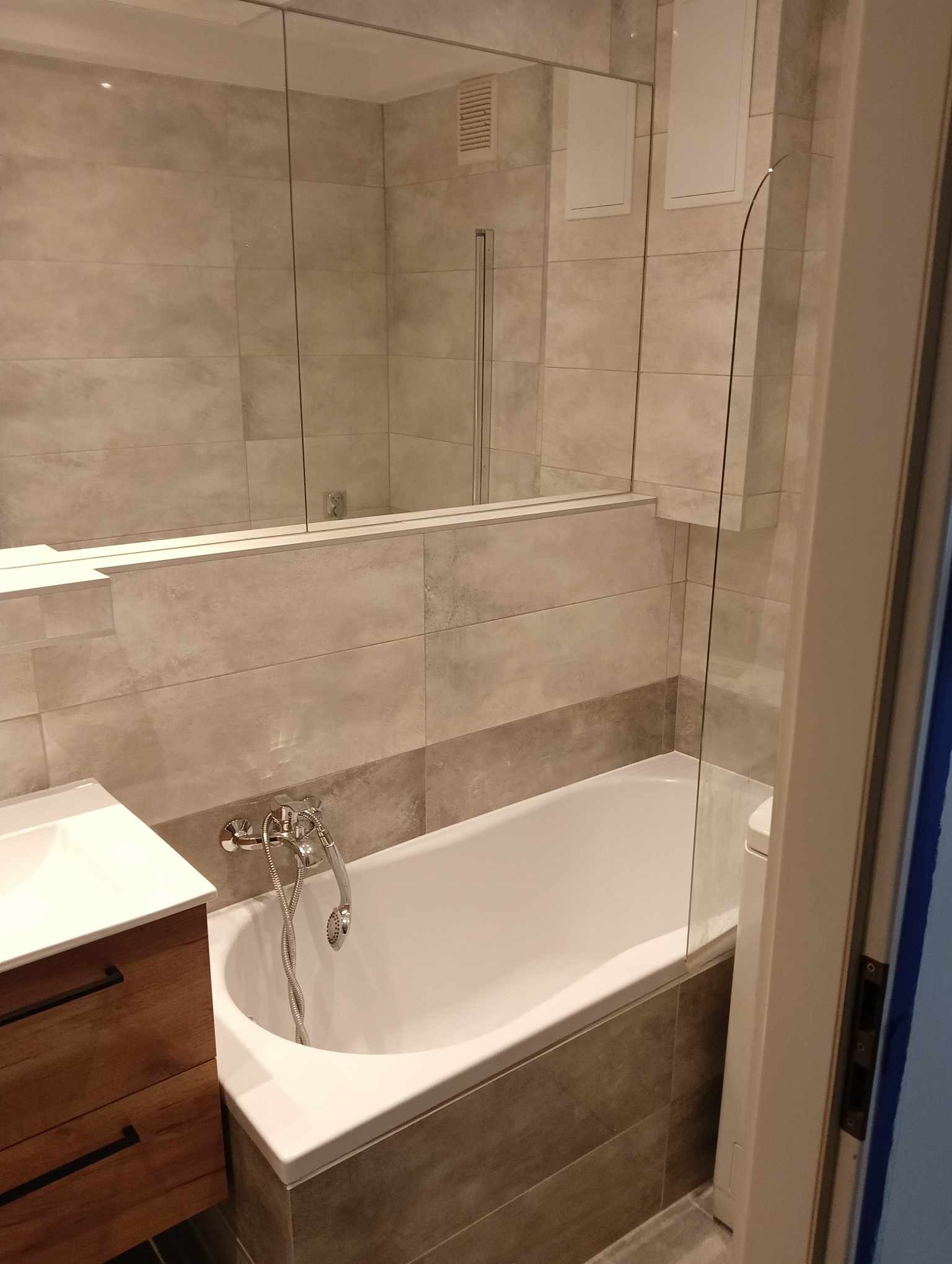 Nowoczesna łazienka z wanną obudowaną szarymi płytkami, szklanym parawanem nawannowym oraz umywalką z szafką z drewnianymi frontami i czarnymi uchwytami. Na ścianach jasnoszare płytki imitujące...