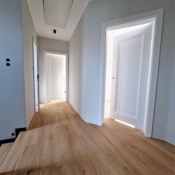 Ambud - Nowoczesny korytarz z panelami podłogowymi w odcieniach drewna, świeżo pomalowane ściany na szaro, białe drzwi z minimalistycznymi ościeżnicami, widoczny właz rewizyjny w suficie.