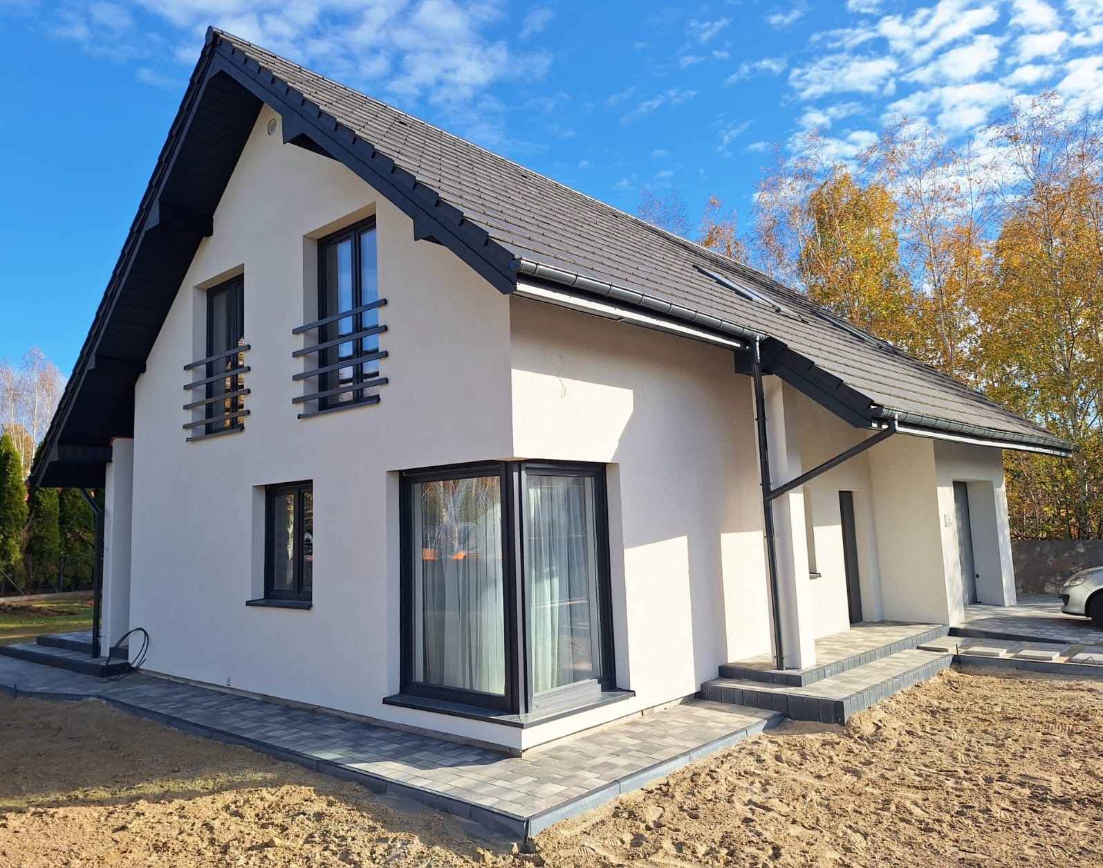 Nowoczesny dom jednorodzinny z szarą dachówką i czarnymi ramami okiennymi, świeżo otynkowany, z chodnikiem z kostki brukowej, w tle drzewa z jesiennymi liśćmi.