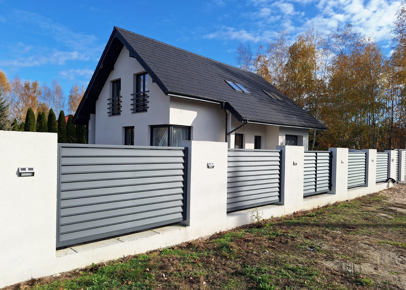 Nowoczesne ogrodzenie panelowe w kolorze szarym z białymi słupkami, oświetleniem i widokiem na dom z ciemną dachówką w tle, jesienny krajobraz.